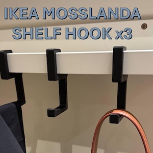 Puede incluir: Ganchos negros para estantes fijados a un estante blanco, con el texto azul "IKEA MOSSLANDA SHELF HOOK x3". Una prenda oscura y un bolso marrón cuelgan de los ganchos. Los ganchos están diseñados para colgar objetos de un estante.