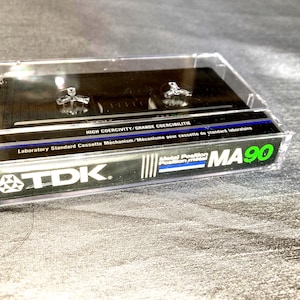 Op de afbeelding: Een TDK MA90 audiocassette in een doorzichtige plastic behuizing. De cassette heeft een zwart interieur met zilveren details. Het label bevat de tekst "HIGH COERCIVITY" en "Laboratory Standard Cassette Mechanism". De behuizing ligt op een grijze, getextureerde ondergrond.