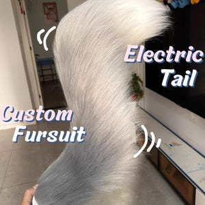 Puede incluir: Una cola de fursuit personalizada con un degradado de piel sintética gris y blanca. La cola es sostenida por una persona. El texto "Electric Tail" y "Custom Fursuit" está en la imagen.