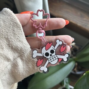 Puede incluir: Un llavero rosa con un diseño de calavera y huesos cruzados con detalles de corazón rojo. El llavero tiene un cierre rosa en forma de flor y un dije de acrílico transparente. El diseño es reminiscente de un tema pirata.