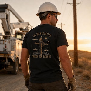 May include: A black short-sleeve t-shirt featuring a graphic on the back of a utility worker sitting on a power pole with the text "THE VIEW IS BETTER WHEN YOU EARN IT" above and below the illustration, along with a sun and mountain scenery.