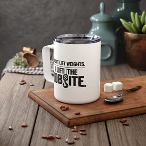Puede incluir: Taza de viaje blanca con tapa y asa negras. La taza tiene el texto "I don't lift weights. I lift the jobsite" en negro. La taza está sobre una tabla de cortar de madera con una cuchara y cubos de azúcar.