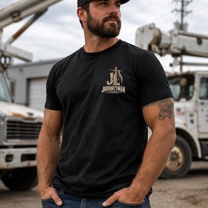 May include: A black short-sleeved t-shirt featuring a graphic on the left chest. The graphic displays the letters JL in a stylized font with a lightning bolt and the word JOURNEYMAN printed underneath in a tan and white color scheme.