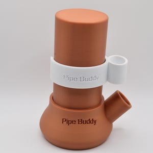 Könnte beinhalten: Eine Wasserpfeife in Terrakotta-Farbe mit einem weißen Band und einem kleinen weißen Halter. Die Worte "Pipe Buddy" sind auf die Pfeife gedruckt. Ein grünes Logo mit einer Cartoon-Figur und den Worten "Pipe Buddy" befindet sich in der unteren rechten Ecke.