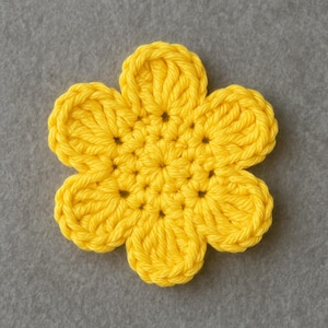 May include: A handmade, bright yellow crochet flower with six petals. The flower is made with a thick yarn, and the stitches are clearly visible. The flower is centered on a gray background, showcasing the vibrant color and texture of the crochet.