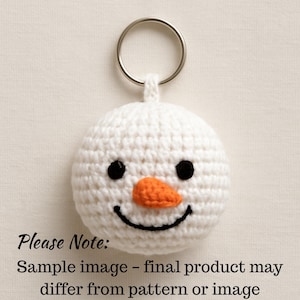 May include: A white crocheted snowman keyring with a silver ring. The snowman features black eyes, an orange carrot nose, and a smiling mouth. The text "Please Note: Sample image - final product may differ from pattern or image" is at the bottom.