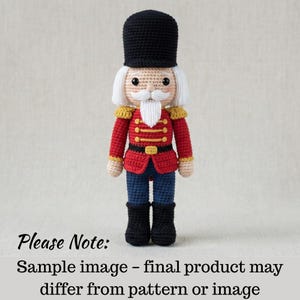 May include: A crocheted nutcracker doll wearing a red jacket with gold detailing, black boots, and a black hat. The doll has a white moustache and hair. The text at the bottom says "Please Note: Sample image - final product may differ from pattern or image."