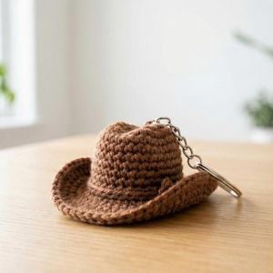 Könnte beinhalten: Ein brauner Cowboyhut-Schlüsselanhänger aus Häkelarbeit mit einer silberfarbenen Kette und einem Schlüsselring. Der Miniaturhut hat ein klassisches Krempen- und Kronen-Design, ideal für Schlüssel oder Taschen.