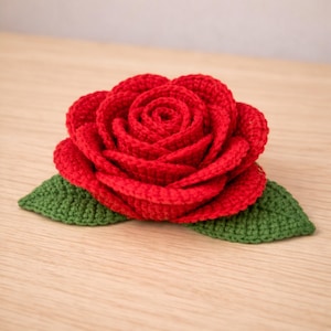 Könnte beinhalten: Eine handgefertigte, rote Häkelrose mit zwei grünen Blättern. Die Rose steht in voller Blüte, mit detaillierten Blütenblättern. Die Blätter sind ein leuchtendes Grün, und die Rose ist auf einer hellen Holzoberfläche platziert. Dieses florale Design ist perfekt für eine Brosche oder einen Dekorationsartikel.