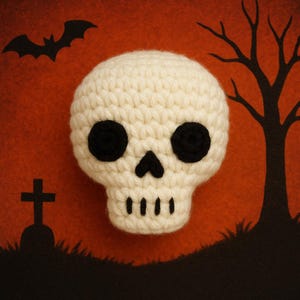 May include: A crocheted skull with black eyes and nose, set against an orange background with a black silhouette of a bat, tree, and tombstone. The skull is a cream color, perfect for Halloween decorations.
