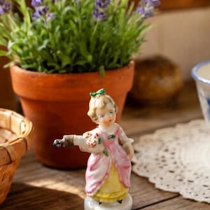 May include: A small porcelain figurine of a girl in a pink and yellow dress, holding a bouquet and a bottle. The figurine has a green bow in her hair. A terracotta pot with lavender plants is in the background.