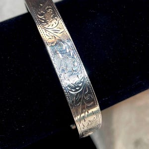 May include: A silver-toned metal bangle bracelet featuring an intricate engraved floral pattern across its entire outer surface, with a polished finish and a thin beaded border running along both edges of the band.