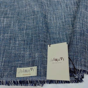 Op de afbeelding: Een blauw-witte geweven linnen sjaal met een gefranjerde rand. De sjaal heeft twee labels, één met de tekst "SOLBIATI WEAVING LINEN CROSSOVER MADE IN ITALY" en de andere met de tekst "SOLBIATI FABRIC MADE IN ITALY".