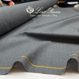 May include: A roll of gray fabric with the text "Loro Piana Super 130s Zelander 280g". The fabric has a subtle texture and a decorative edge with yellow and red stripes.