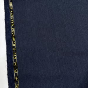 May include: Close-up of navy blue fabric with a subtle texture. A gold border on the left side features the text "HIGH TWISTED ZELANDER 3 PLY CLOT". The fabric appears to be a high-quality material, possibly for suiting or dressmaking.