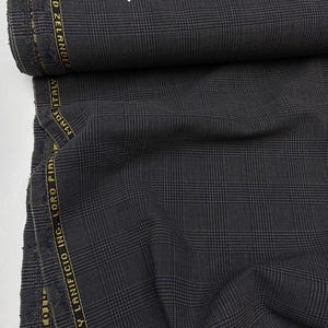May include: A roll of dark gray fabric with a plaid pattern. The fabric has a gold-colored stripe with text that reads "TWISTED ZELANDER" and "MADE IN ITALY". The Loro Piana logo is visible.