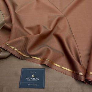 Op de afbeelding: Een gevouwen stuk bruine stof met een subtiele glans. De stof heeft een fijne textuur en is voorzien van het label "SCABAL FINEST CLOTH". Gouden randen met tekst en een logo zijn zichtbaar langs de randen. De stof lijkt van hoge kwaliteit te zijn.