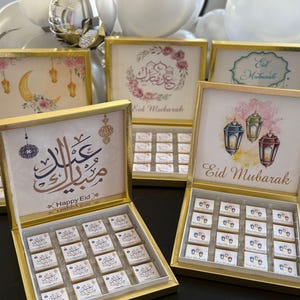 May include: Gold-framed boxes with decorative lids and chocolates inside. The lids feature watercolor illustrations of lanterns, crescent moons, and floral arrangements, with the text "Eid Mubarak" in English and Arabic calligraphy. The boxes are open, revealing rows of chocolates.