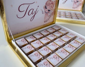 May include: A gold-colored box with a hinged lid, filled with small, square chocolates. The lid features the name "Taj" in a script font, along with an illustration of a girl. Each chocolate is individually wrapped with the same design.