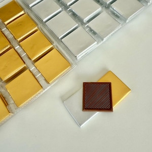 May include: Overhead view of square chocolates in gold and silver foil wrappers, arranged in clear plastic trays. One unwrapped chocolate square with a ridged surface sits atop a silver wrapper, next to a gold wrapper.