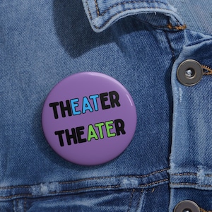 May include: A round, purple button with the word "THEATER" in black, blue, and green text. The button is pinned to a blue denim jacket.