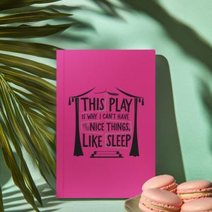 Op de afbeelding: Een felroze notitieboek met de zwarte tekst "THIS PLAY IS WHY I CAN'T HAVE NICE THINGS, LIKE SLEEP". Daarnaast een bord met roze macarons. De achtergrond is licht turquoise met schaduwen van palmbladeren.
