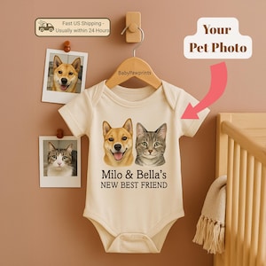 May include: A cream-colored baby onesie featuring illustrations of a dog and a cat, with the text "Milo & Bella's NEW BEST FRIEND." The onesie is hanging on a wooden hanger. Two pet photos are pinned above the onesie.