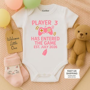 May include: White babygrow with pink text: "PLAYER 3 HAS ENTERED THE GAME EST. JULY 2026." The design includes a pink games controller and a baby bottle icon. A teddy bear and baby socks are also in the image.