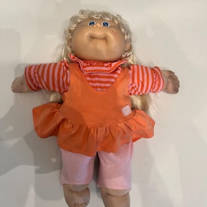 May include: A vintage Cabbage Patch Kid doll with blonde hair and blue eyes. The doll is wearing an orange top with a ruffled skirt and pink and orange striped sleeves. The doll is also wearing pink ribbed pants.