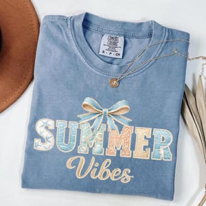 May include: A blue Comfort Colors t-shirt with the embroidered words "SUMMER VIBES" in a decorative font. The word "SUMMER" has a bow above it. A gold necklace with a pendant is on the shirt.