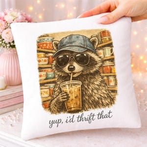 May include: White square pillow with a raccoon wearing sunglasses and a baseball cap, sipping an iced coffee. The raccoon is in front of shelves of mugs and bowls. The text "yup, i'd thrift that" is printed on the pillow.