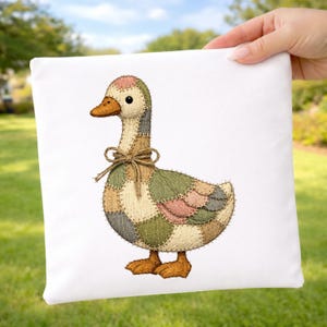 May include: A decorative square pillow featuring a patchwork goose design. The goose has a brown beak and feet, with a beige, green, and pink patchwork body. A brown twine bow adorns the goose's neck. The pillow is held against a background of green grass and trees.