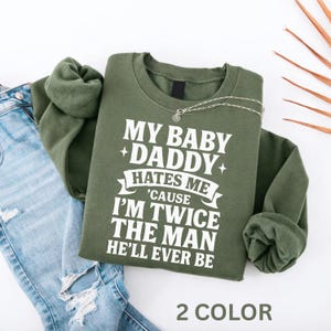 Puede incluir: Una sudadera verde oliva con el texto "MY BABY +DADDY+ HATES ME 'CAUSE I'M TWICE THE MAN HE'LL EVER BE" en blanco. Un collar plateado está drapeado en la parte delantera. Las palabras "2 COLOR" están en la parte inferior. Se ven unos vaqueros azules.