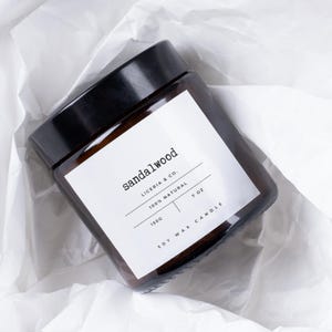 Könnte beinhalten: Ein quadratisches braunes Glas-Kerzenglas mit schwarzem Deckel, beschriftet mit "sandalwood". Das weiße Etikett lautet ebenfalls "LICERIA & CO.", "100% NATURAL" und "7 oz". Die Kerze ist in weißem Seidenpapier verpackt und enthält 150g Sojawachs.