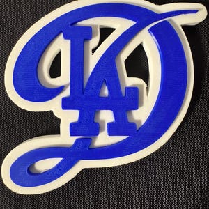 Puede incluir: Un llavero blanco y azul con el logotipo de los Dodgers de Los Ángeles. El logotipo es un "LA" estilizado en azul, delineado en blanco. El llavero tiene un anillo de metal para sujetar.