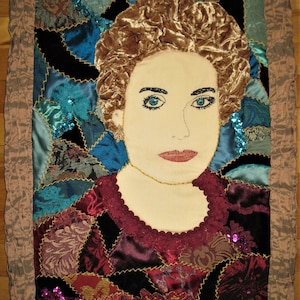 May include: A quilted portrait of a woman with brown hair and blue eyes. The portrait is made of various fabrics in shades of blue, red, and gold. The woman is wearing a burgundy dress with a lace neckline.