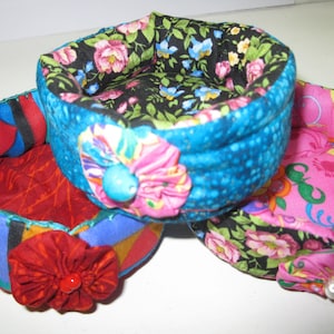 May include: Three fabric storage baskets with floral patterns. The baskets are made of colorful fabric and have a round shape. The baskets are decorated with fabric flowers.