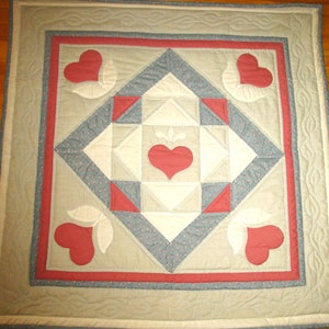 May include: A quilt with a patchwork design featuring a central heart and four corner hearts. The quilt is made with a variety of fabrics in shades of pink, grey, and white.