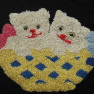 May include: A yellow, blue, and pink felt appliqué of two white kittens in a basket. The kittens have black eyes and red noses.