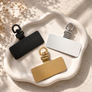 May include: Three rectangular metal tags with rounded corners, each featuring a swivel lobster clasp attachment at the top. The set includes one matte black tag, one silver-toned tag, and one brushed gold-toned tag, all arranged in a cluster.