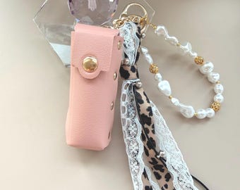 May include: A pink leather case with a gold button closure, attached to a keychain with a leopard print ribbon, white lace, and a pearl and gold bead bracelet. A clear crystal prism sits behind the case.