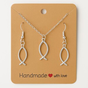 Può includere: Un set di gioielli color argento composto da una collana e due orecchini, ciascuno con un ciondolo a forma di pesce. Il set è presentato su una carta marrone con la scritta "Handmade with love".