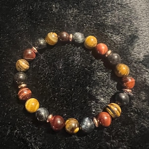 May include: A beaded bracelet featuring a variety of natural stone beads in shades of brown, yellow, and gray. The bracelet includes copper-colored spacers and is displayed on a dark, textured surface.