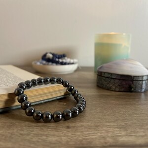 May include: A dark gray beaded bracelet rests on a wooden surface next to an open book. A small, decorative shell-shaped box and a candle are in the background. The bracelet is made of smooth, round beads.