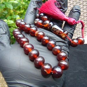 Puede incluir: Un collar de cuentas de oración de ámbar oscuro con una borla roja. Las cuentas son redondas y pulidas, con un color marrón rojizo intenso. La borla está hecha de hilos rojos gruesos y retorcidos.