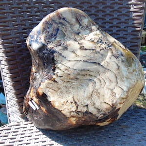 Puede incluir: Una piedra grande, de forma irregular, con una superficie pulida. La piedra presenta una mezcla de colores beige, marrón y marrón oscuro, con un aspecto texturizado y en capas. Está sobre una silla de mimbre marrón.
