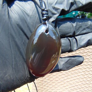 Op de afbeelding: Een donkere, ovale hanger met een glanzend oppervlak, bevestigd aan een zwart koord. Het oppervlak van de hanger reflecteert het omgevingslicht. De hanger wordt gepresenteerd tegen een donkere achtergrond.