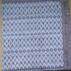 May include: A rectangular textile with a repeating pattern of gray diamonds and floral motifs on a light blue background. The edges are bordered with a dark gray trim. A section on the right features a textured design and text.