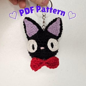 May include: A black crocheted cat keychain with large white eyes, light purple inner ears, a small pink nose, and a bright red bow tie. The text PDF Pattern is displayed above the cat, flanked by two purple heart outlines on each side.