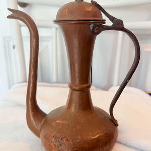 May include: A copper pitcher with a long, curved spout, a rounded base, and a tall, slender neck. It features a domed lid with a small knob handle and a dark, curved metal handle attached to the side, showing a weathered, metallic finish.